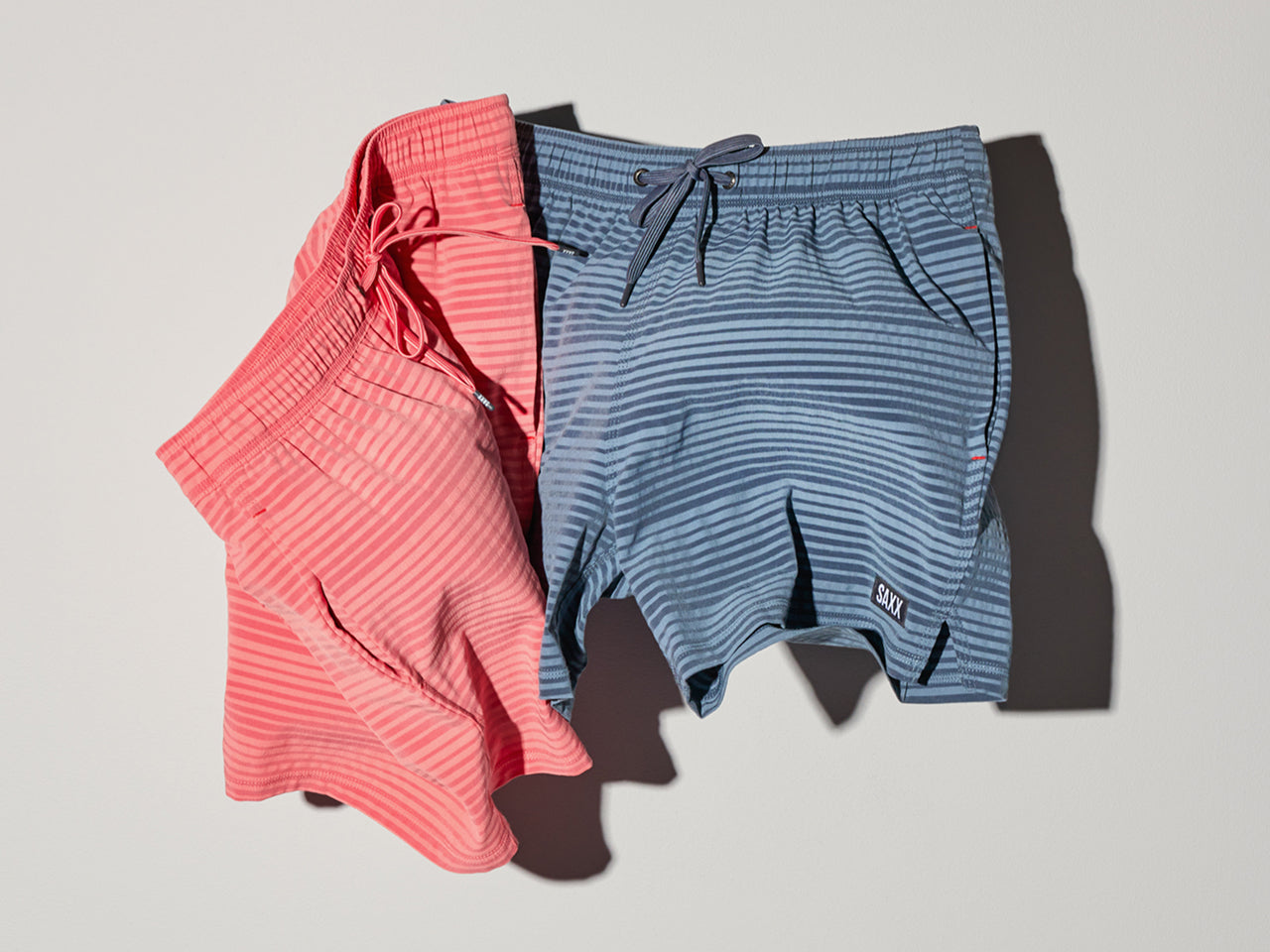 Two pairs of striped swim shorts in tonal coral and tonal blue colors set against a grey background.