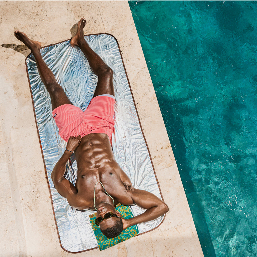 A man with a shredded athletic physique laying next to a pool. He is wearing pink swim shorts.