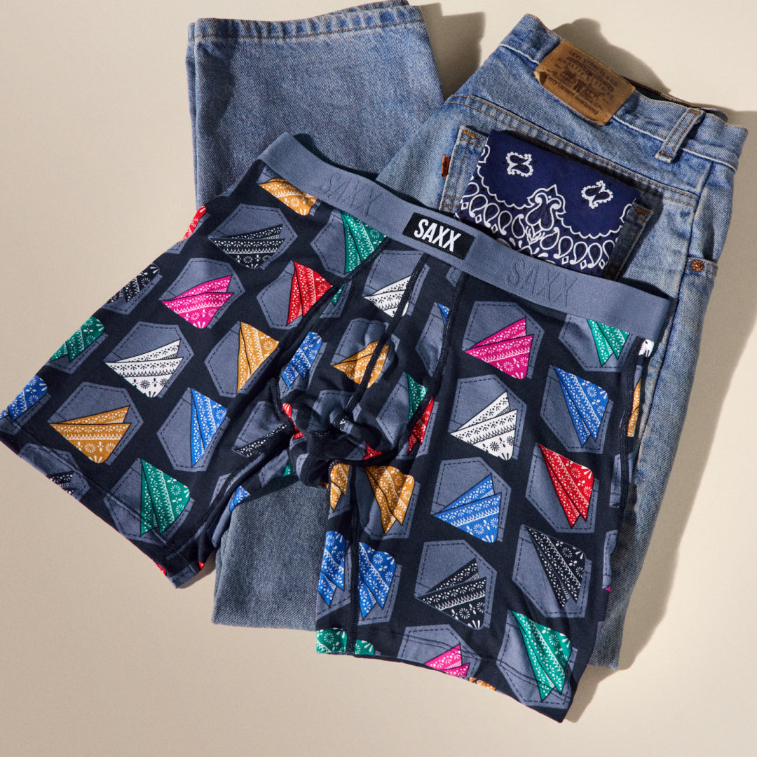 Stack of SAXX boxer briefs with geometric pattern and a pair of jeans on a beige background