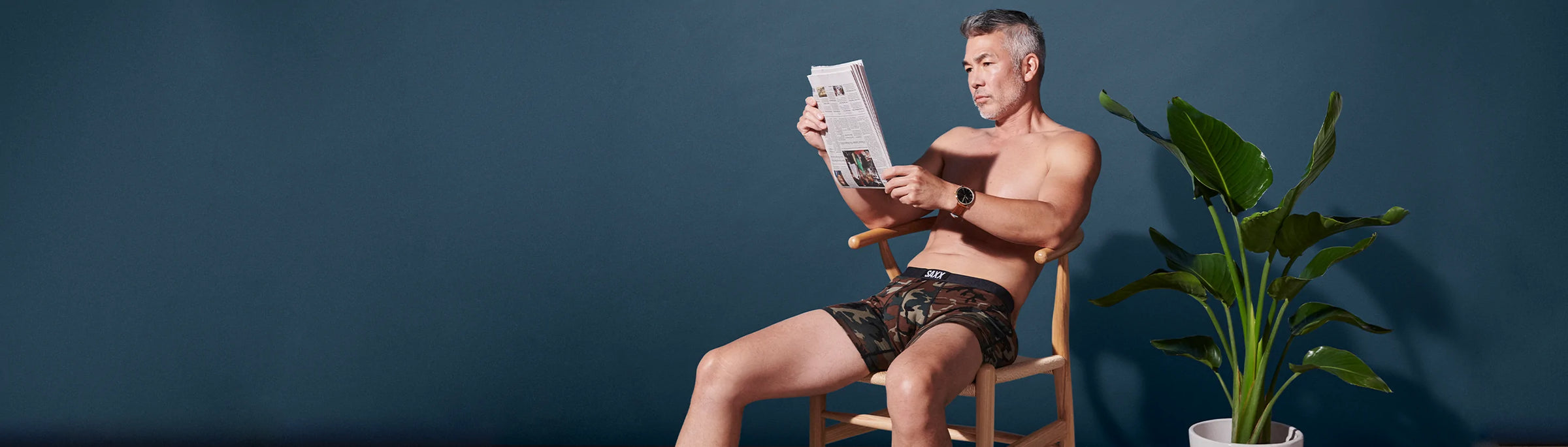 A person sitting in a chair reading the newspaper while wearing the Vibe Super Soft Boxer Brief in Woodland Camo