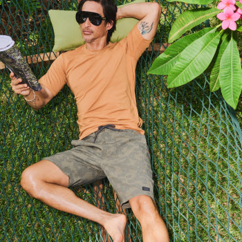 Man in orange shirt and camo shorts reading magazine in hammock