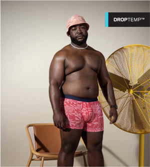 A man wearing a bucket hat and a pair of pink tonal patterned boxer briefs standing in front of a wood chair and a large yellow electric fan. 