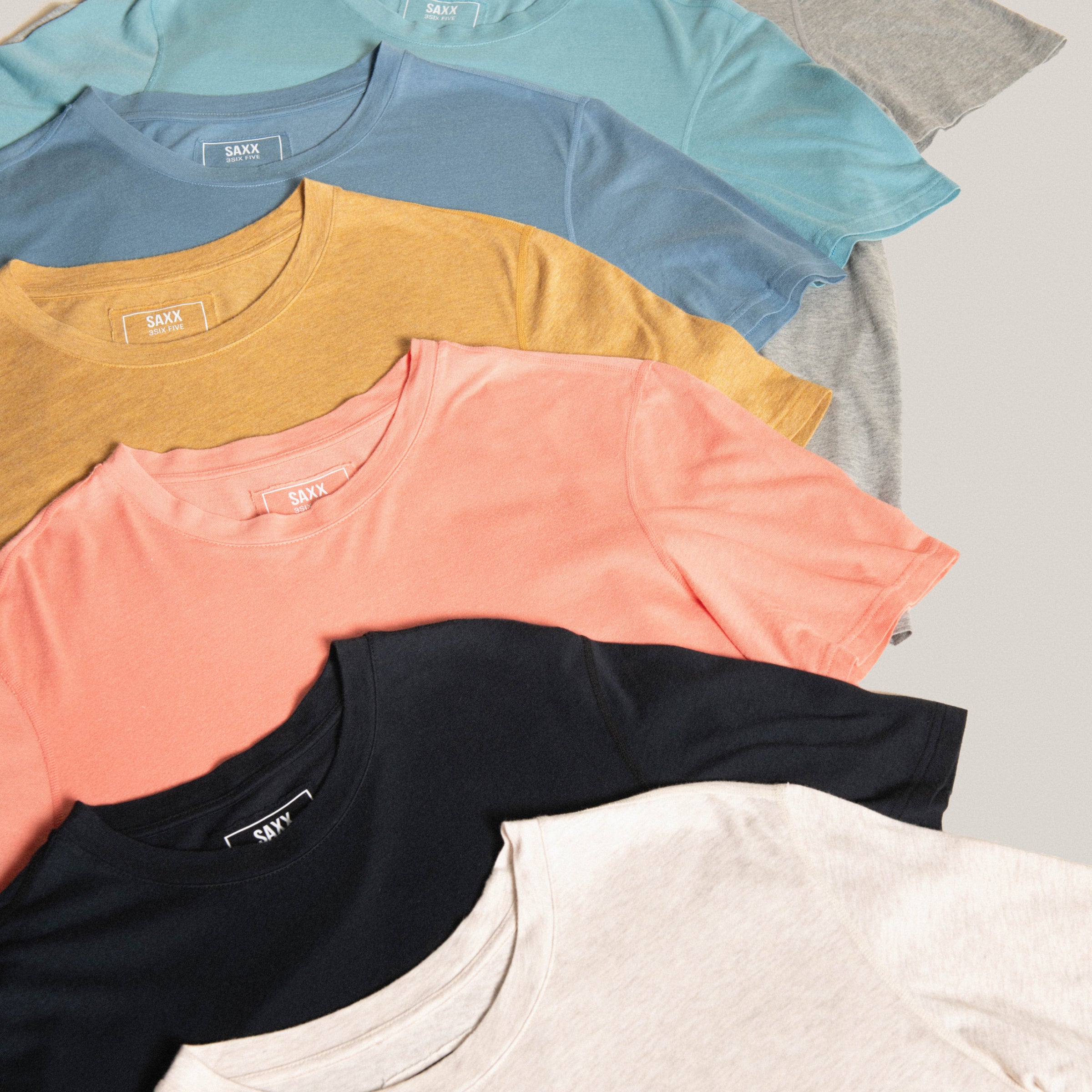 A variety of solid color SAXX short sleeve crew neck shirts laid out over a neutral background.