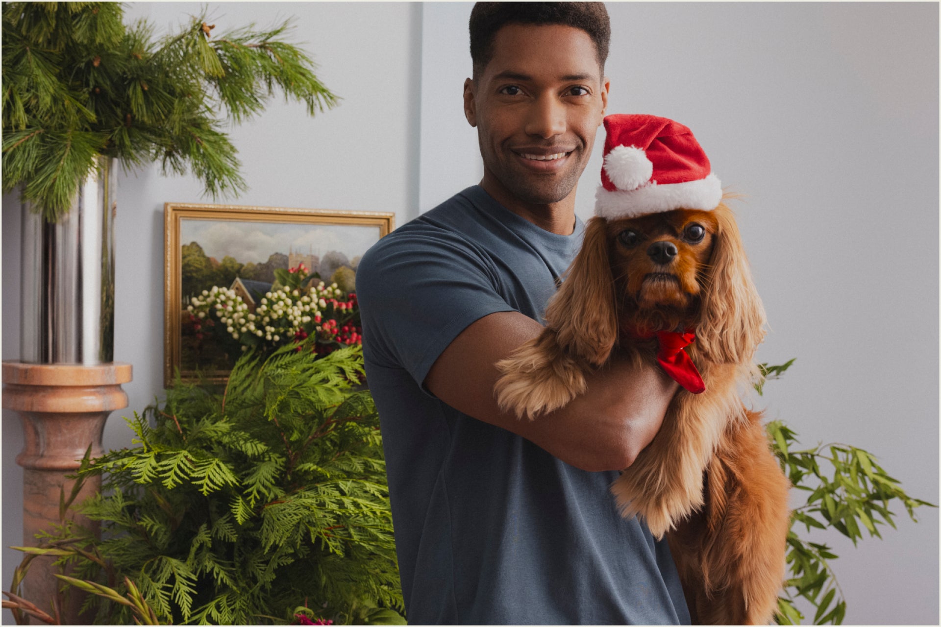 A man holding a dog. The dog wears a Santa Claus hat.