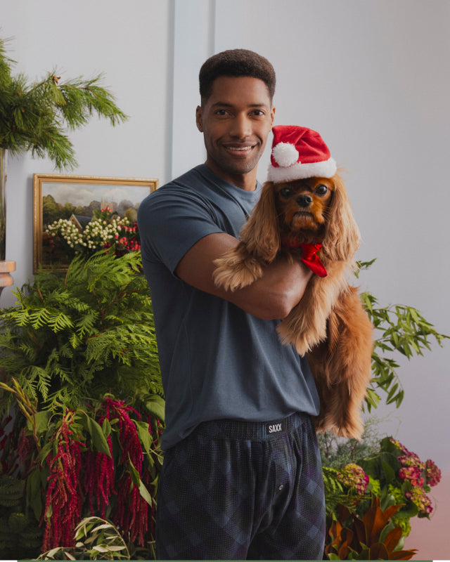 A man holding a dog. The dog wears a Santa Claus hat.