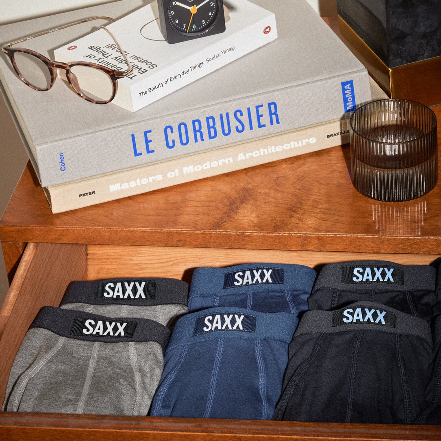 Neatly folded boxer briefs seen in an open top drawer of a dresser with books on top.