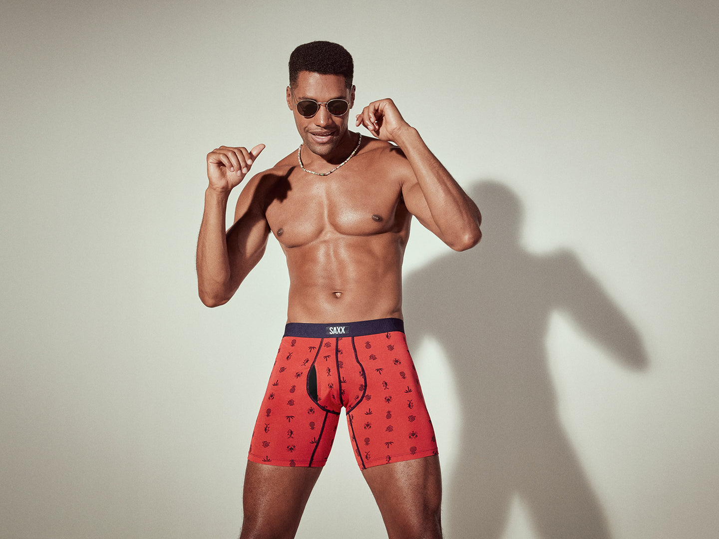 A man standing against a beige background. He wears red boxer briefs and sunglasses. His arms are raised.