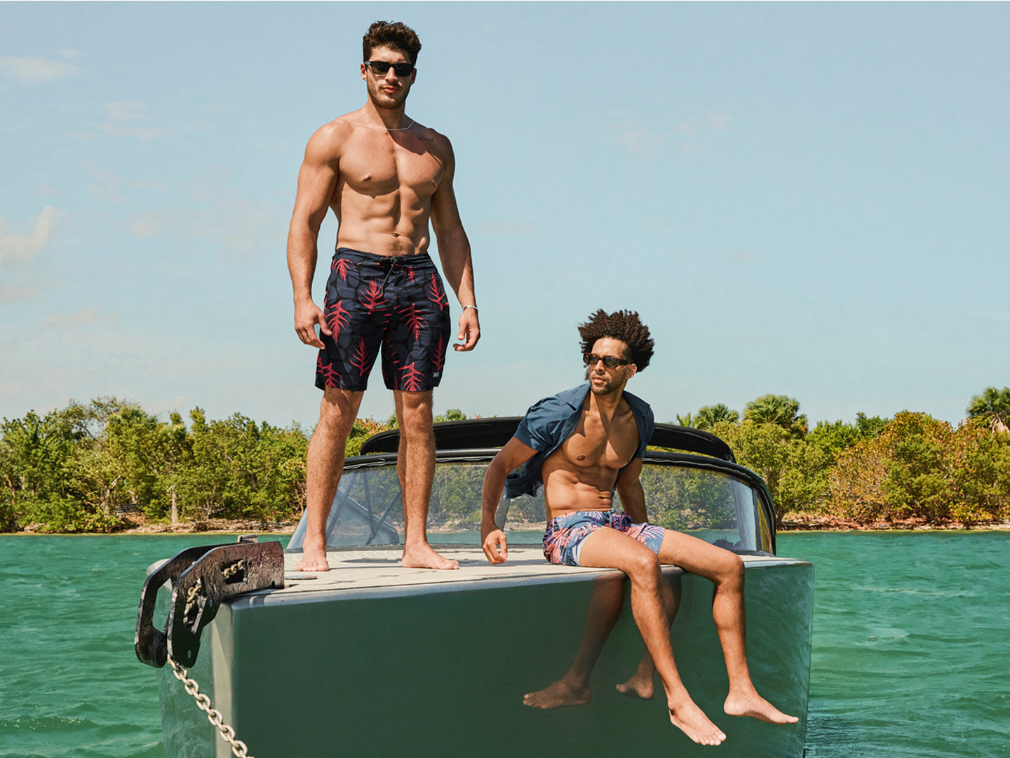 Two men on the hull of a boat wearing swim shorts. The man on the left is standing while the man on the right hands his legs over the edge.