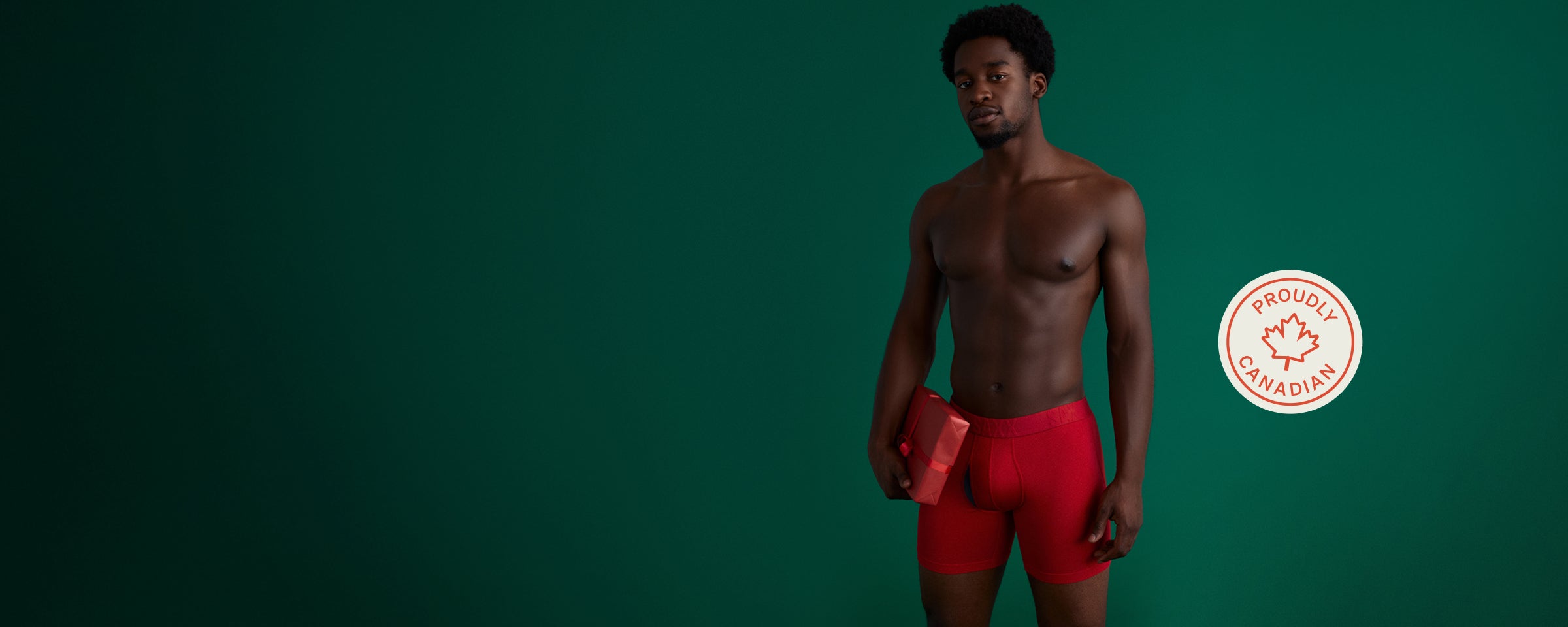 A person wearing red SAXX Boxer Briefs holding a red present, with a branded sticker that says Proudly Canadian