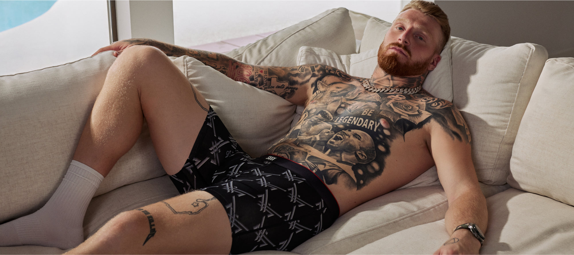 Man with tattoos lying on a couch wearing black boxer briefs.