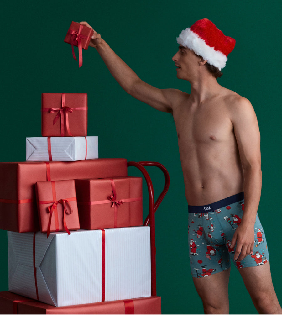 Shirtless man in underwear and Santa hat stacking holiday gifts