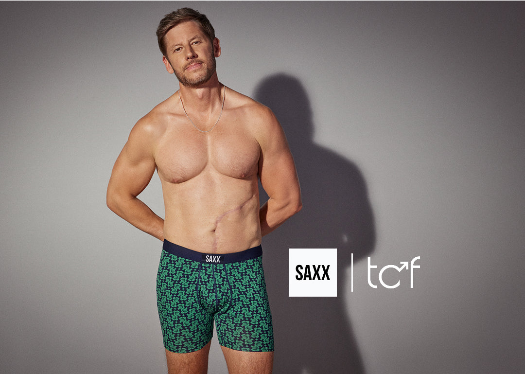 A man standing against a grey background. He is wearing a pair of boxer briefs in a green and blue geometric print. 
