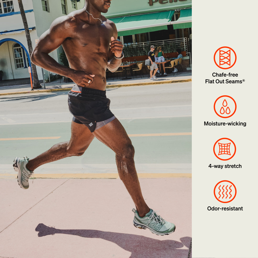 Man jogging on sidewalk in black SAXX Performance shorts. Icons for Flat Out Seams®, moisture-wicking, 4-way stretch, odor-resistant.