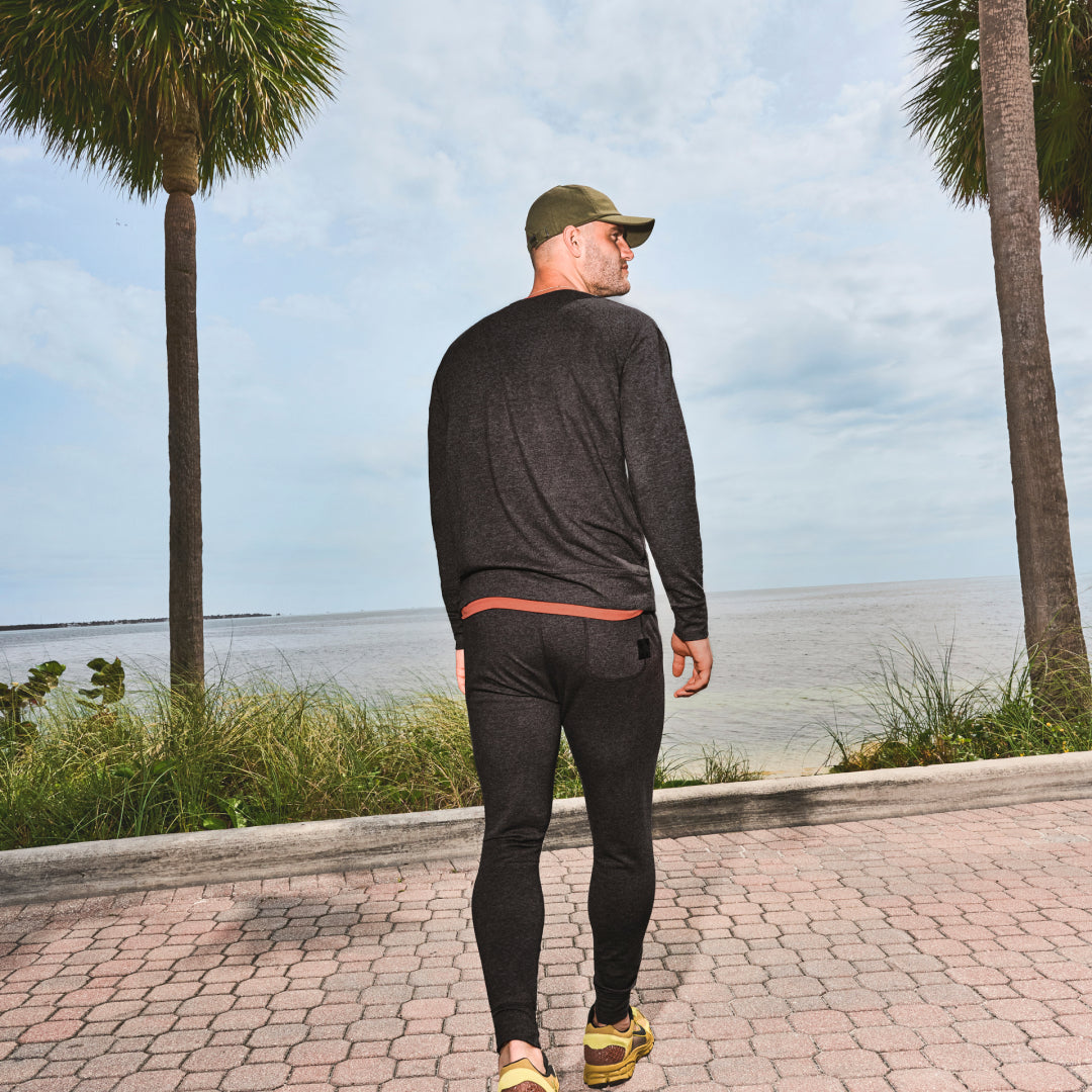 A man walking away from camera in grey SAXX activewear set mid-stride toward ocean and palm trees.