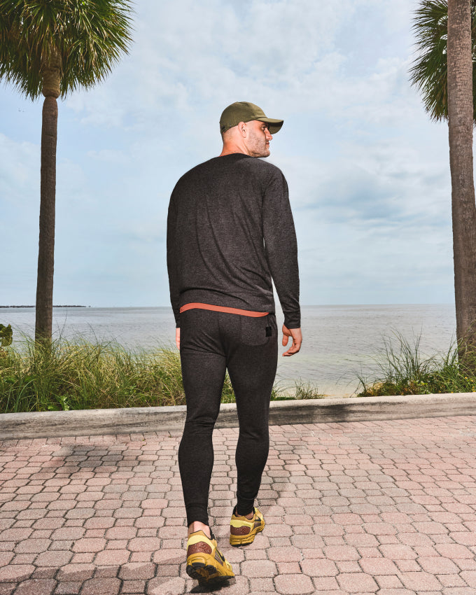 A man walking away from camera in grey SAXX loungewear set mid-stride toward ocean and palm trees.