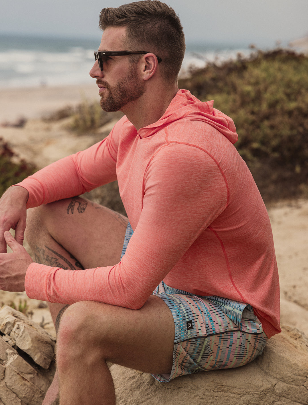 Man wearing a pink hoodie and patterned shorts sitting on a rock by the beach.
