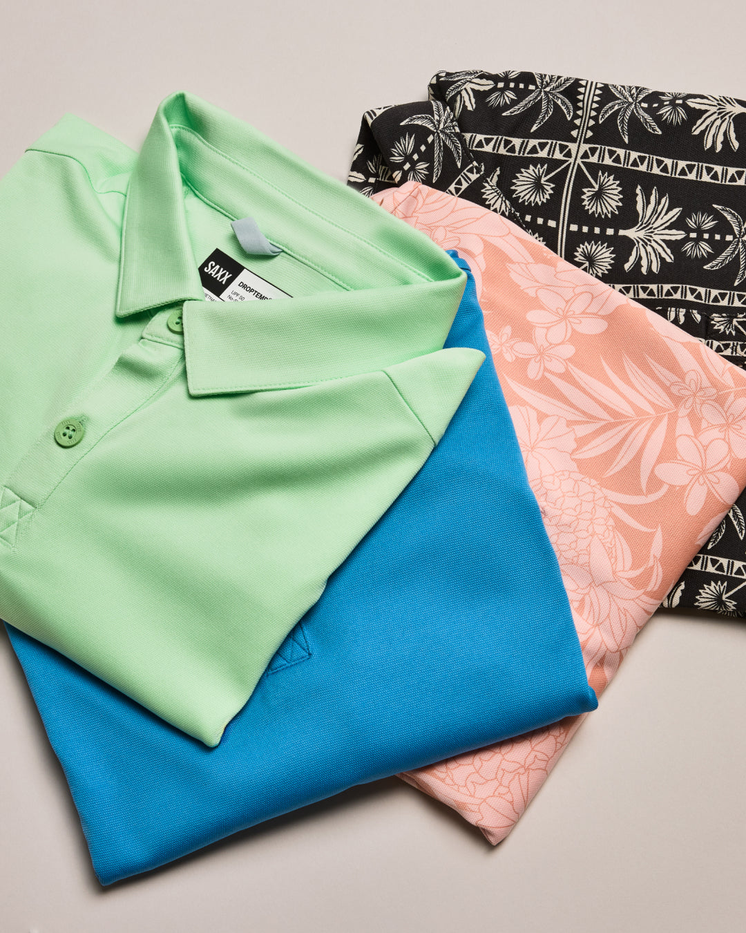 Folded shirts in green, blue, and pink with a patterned shirt in the background on a light gray surface.