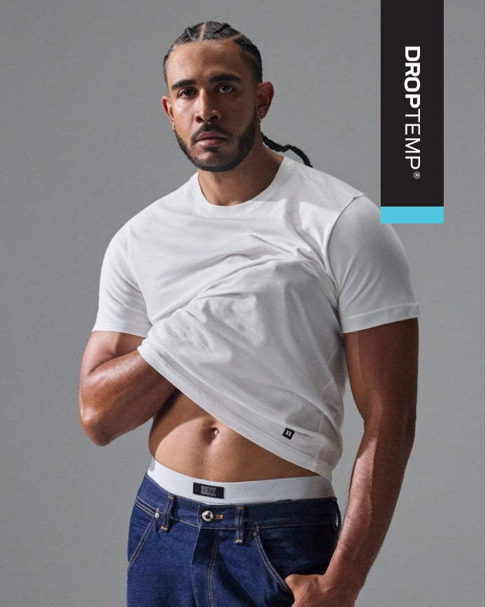 A muscular man wearing a white short-sleeve crop top t-shirt by DROPTEMP and blue jeans, posing against a gray studio background with a vertical DROPTEMP brand banner on the right.