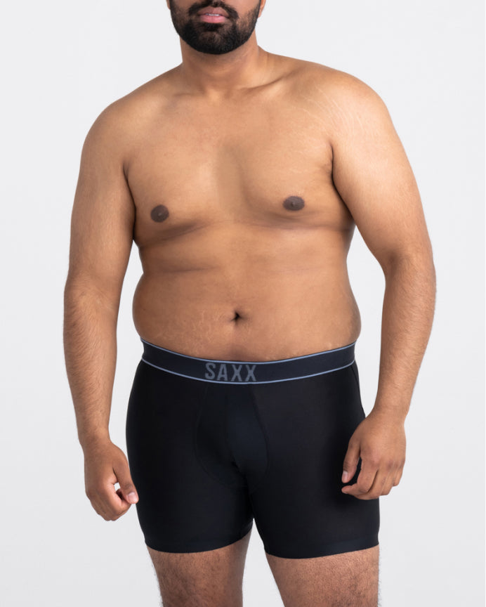 A man wearing SAXX black boxer briefs with a dark gray elastic waistband, shown from the waist down against a light gray background.