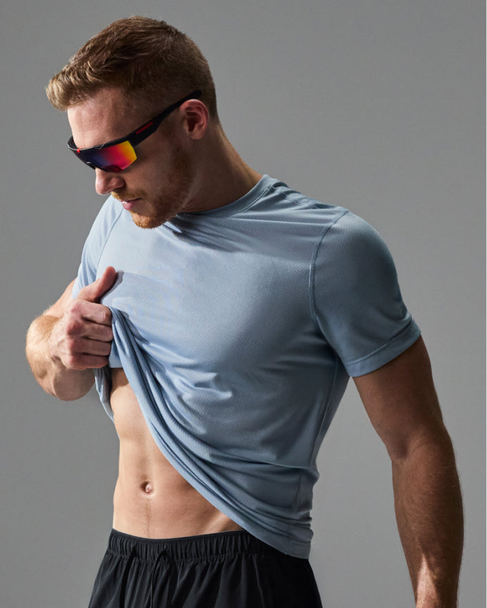A man wearing a light blue athletic t-shirt and black shorts, lifting the shirt to show its fit and texture. He is wearing black sunglasses with colorful mirrored lenses against a solid gray studio background.