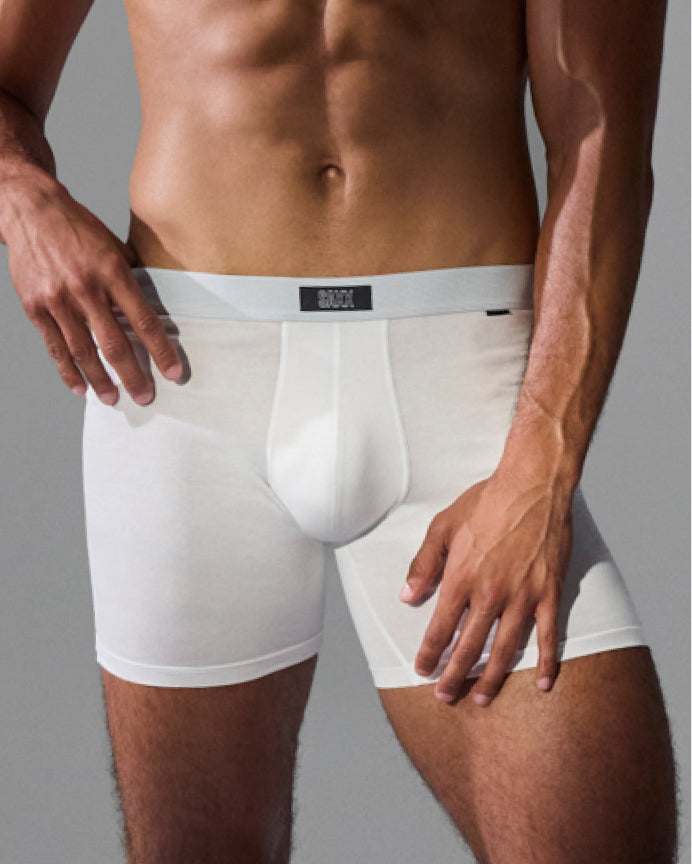 A close-up of a male model wearing white boxer briefs against a plain gray studio background. The waistband has a small black brand label reading BRAVO.