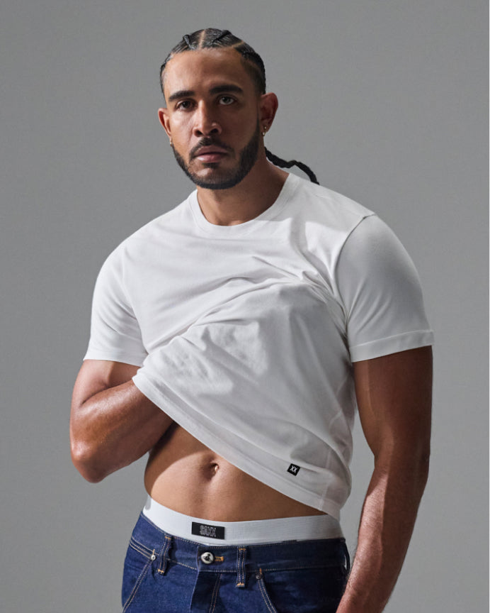 A muscular man wearing a white short-sleeve crewneck t-shirt and blue jeans, lifting the shirt up to show the elastic hem and inner waistband. The brand name 'G-1000' is visible on the inner label.