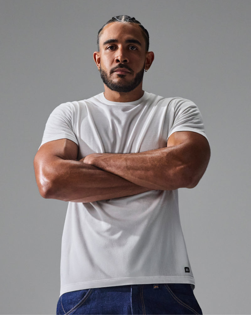 A muscular man with braided hair and a beard wearing a white short-sleeve crewneck t-shirt and blue jeans, standing with his arms crossed against a solid gray studio background.