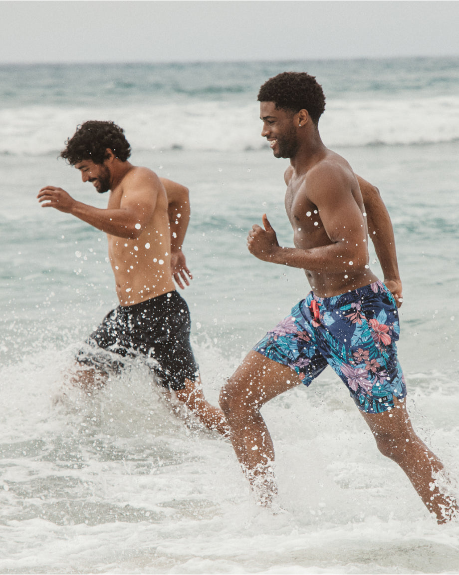 Two men running in the ocean with one wearing colorful SAXX swim trunks.