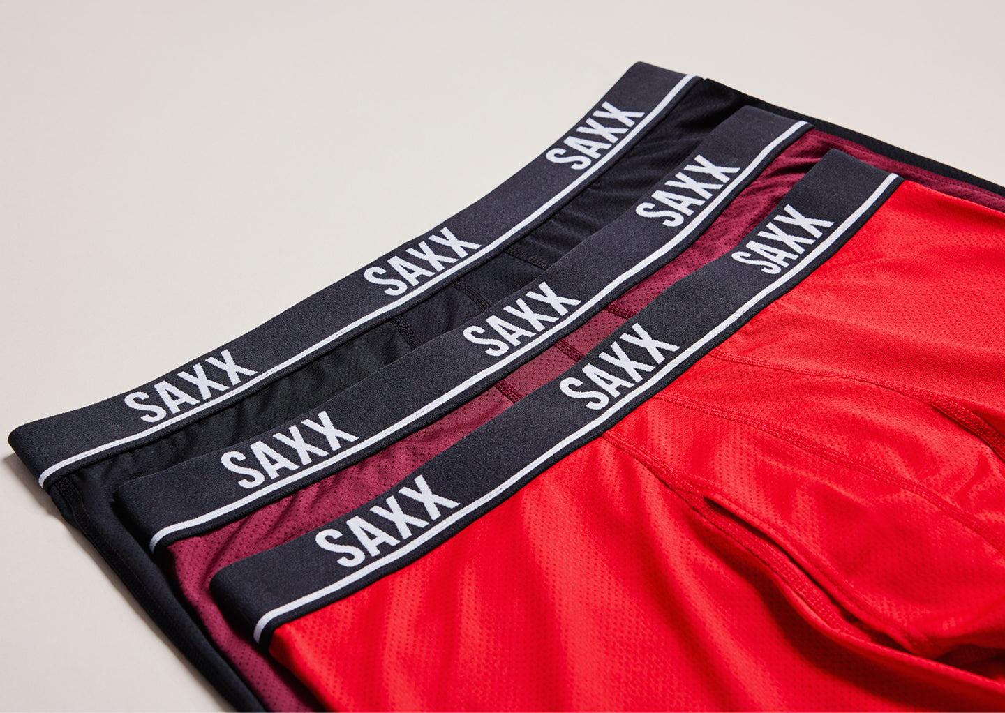 Three SAXX boxer briefs in red and black, with black waistbands on a light plain background.