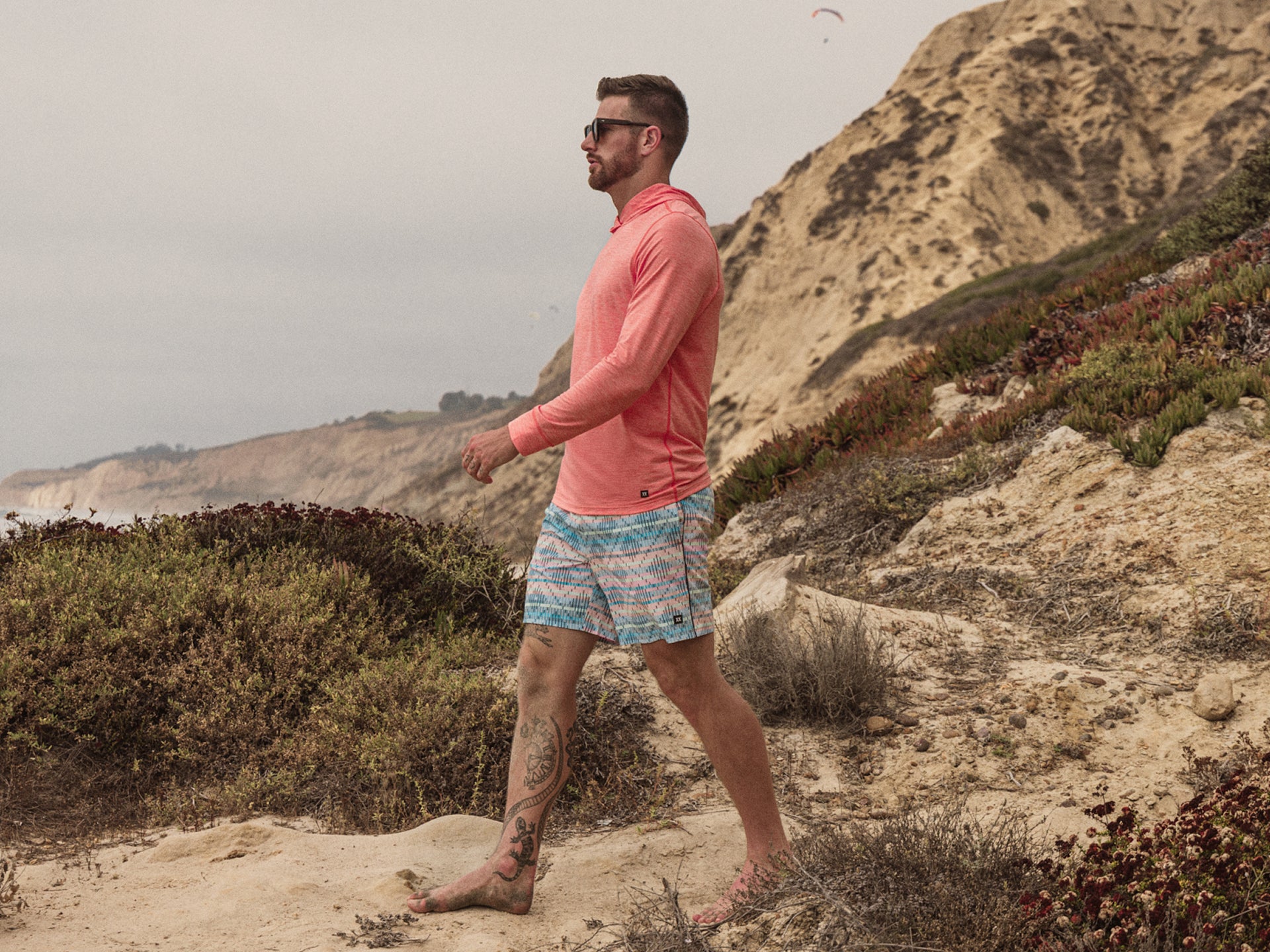 Man in SAXX DropTemp Hoodie and Oh Buoy Swim Shorts walking along a beach