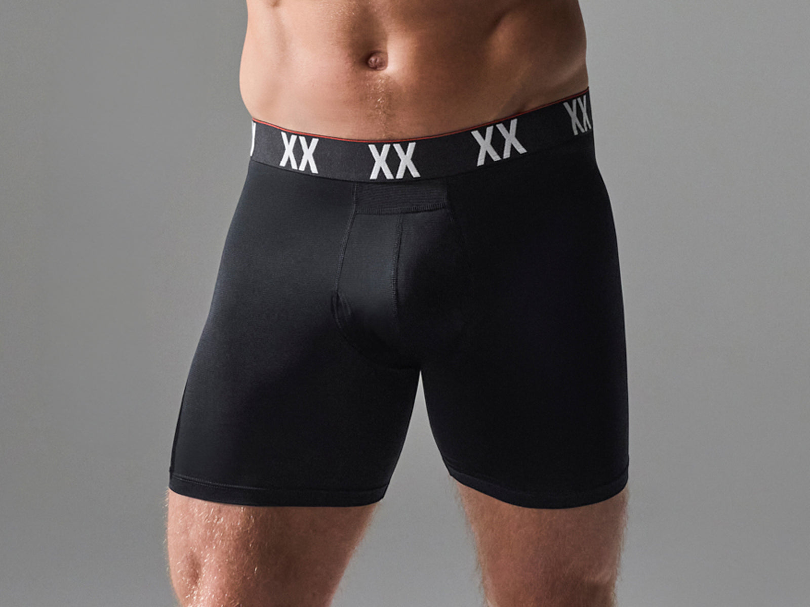 A close-up of a male model wearing black boxer briefs with a black waistband featuring white 'XX' logos, set against a solid gray studio background.