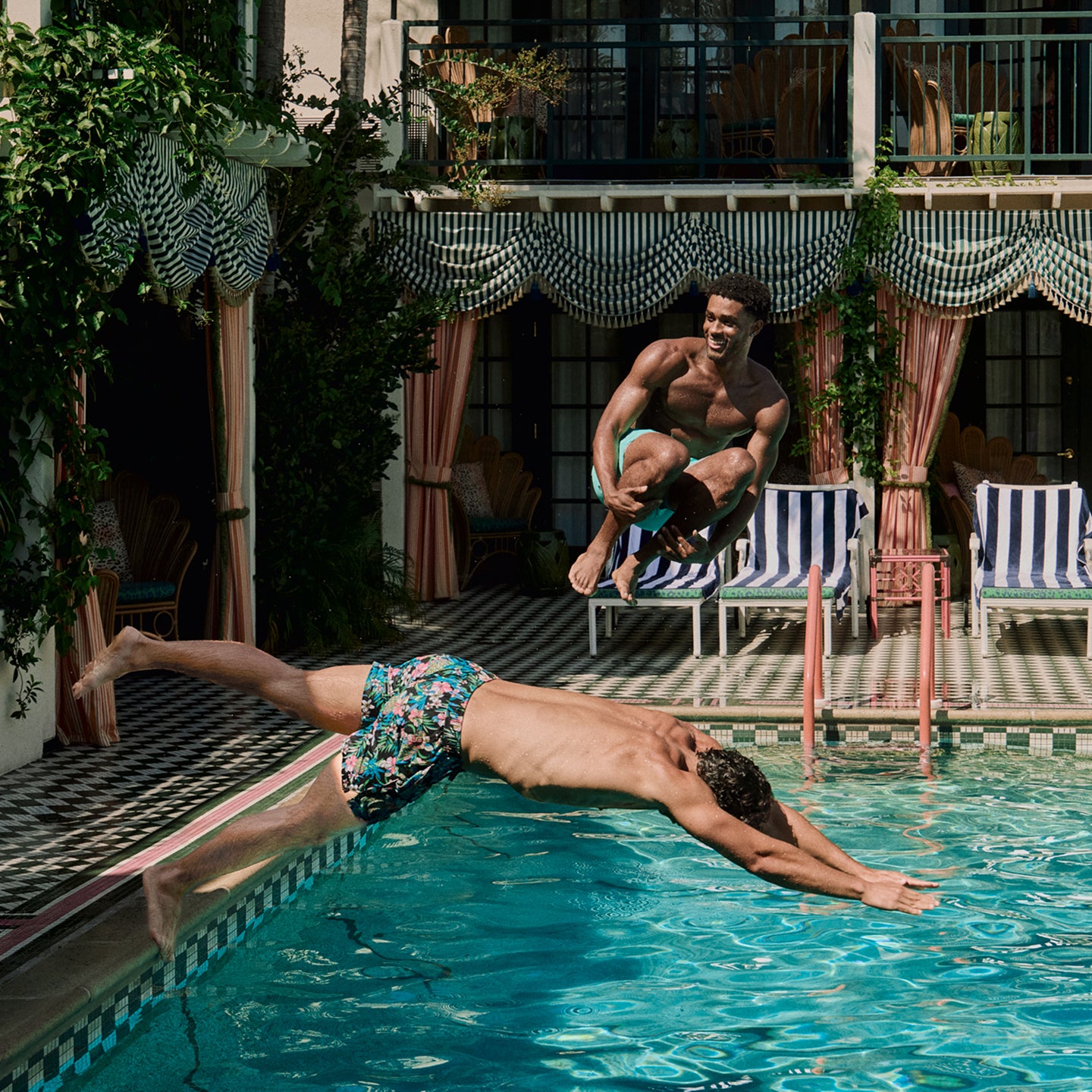 Two men diving into a pool with lounge chairs and plants in the background