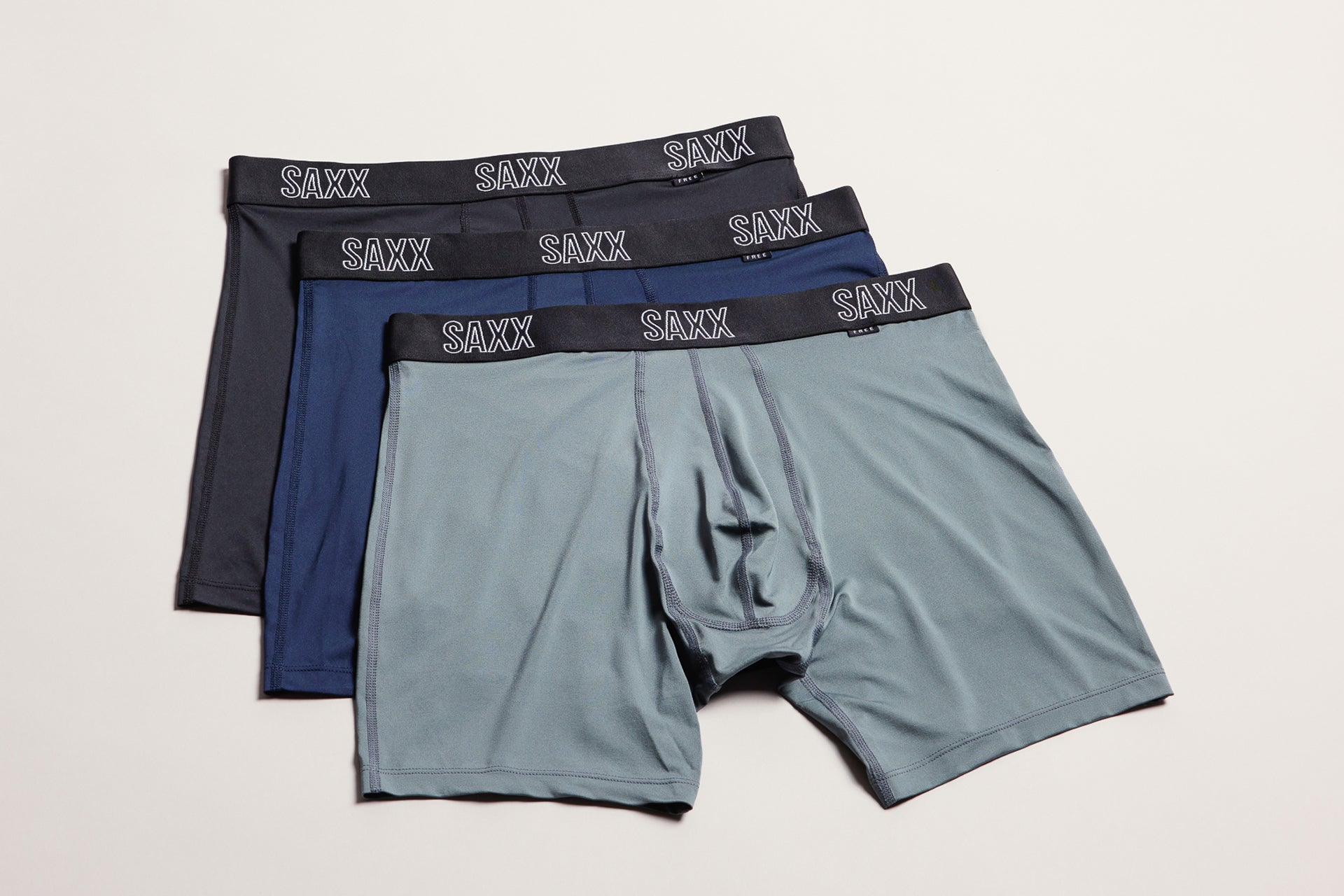 Four pairs of SAXX men's boxer briefs in dark gray, navy blue, and light gray, arranged in a fan shape on a light beige background.