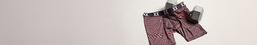 A pair of men's boxer briefs with a red and black abstract pattern and a black waistband featuring white 'XXL' text, shown next to a gray box on a light beige background.