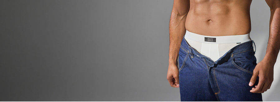 A man's torso wearing an open pair of blue jeans with a white boxer brief underneath, shown against a plain gray studio background.