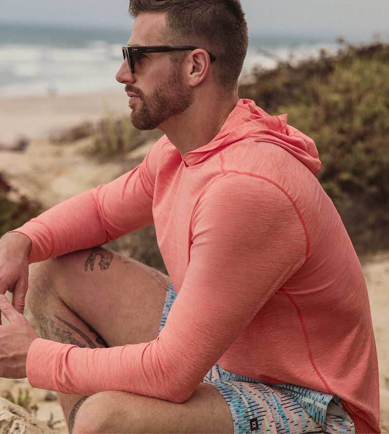 Man wearing lightweight coral hooded top sitting on a trail overlooking the beach