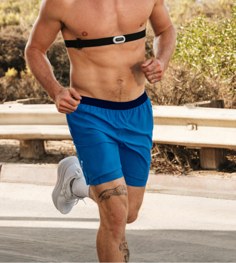 Man running in blue shorts with no shirt and heart rate monitor along coastal trail