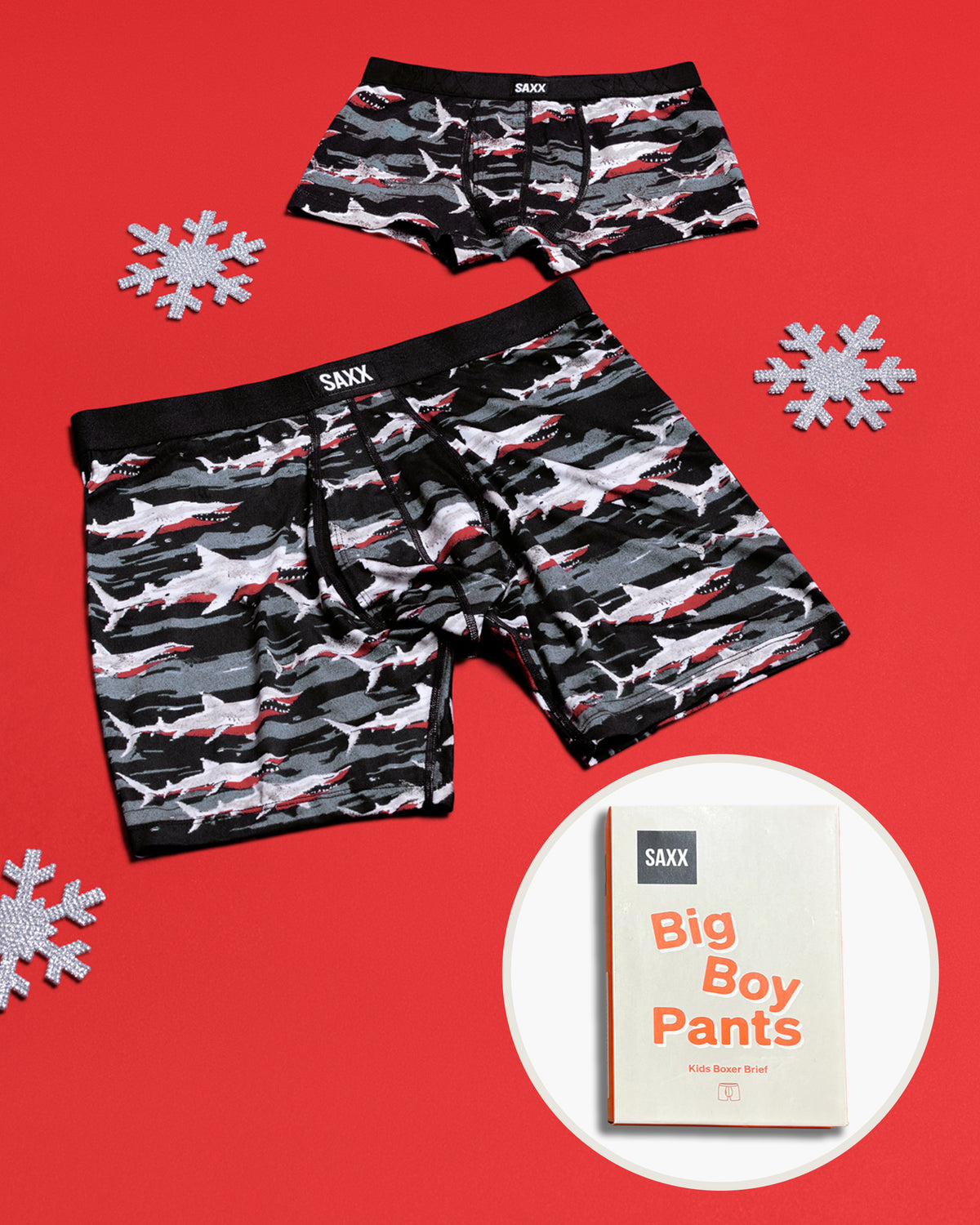 Two pairs of SAXX Father/Son boxer briefs with a shark camouflage pattern in black, gray, and red, displayed on a red background with a circular inset showing a box with 'Big Boy Pants'