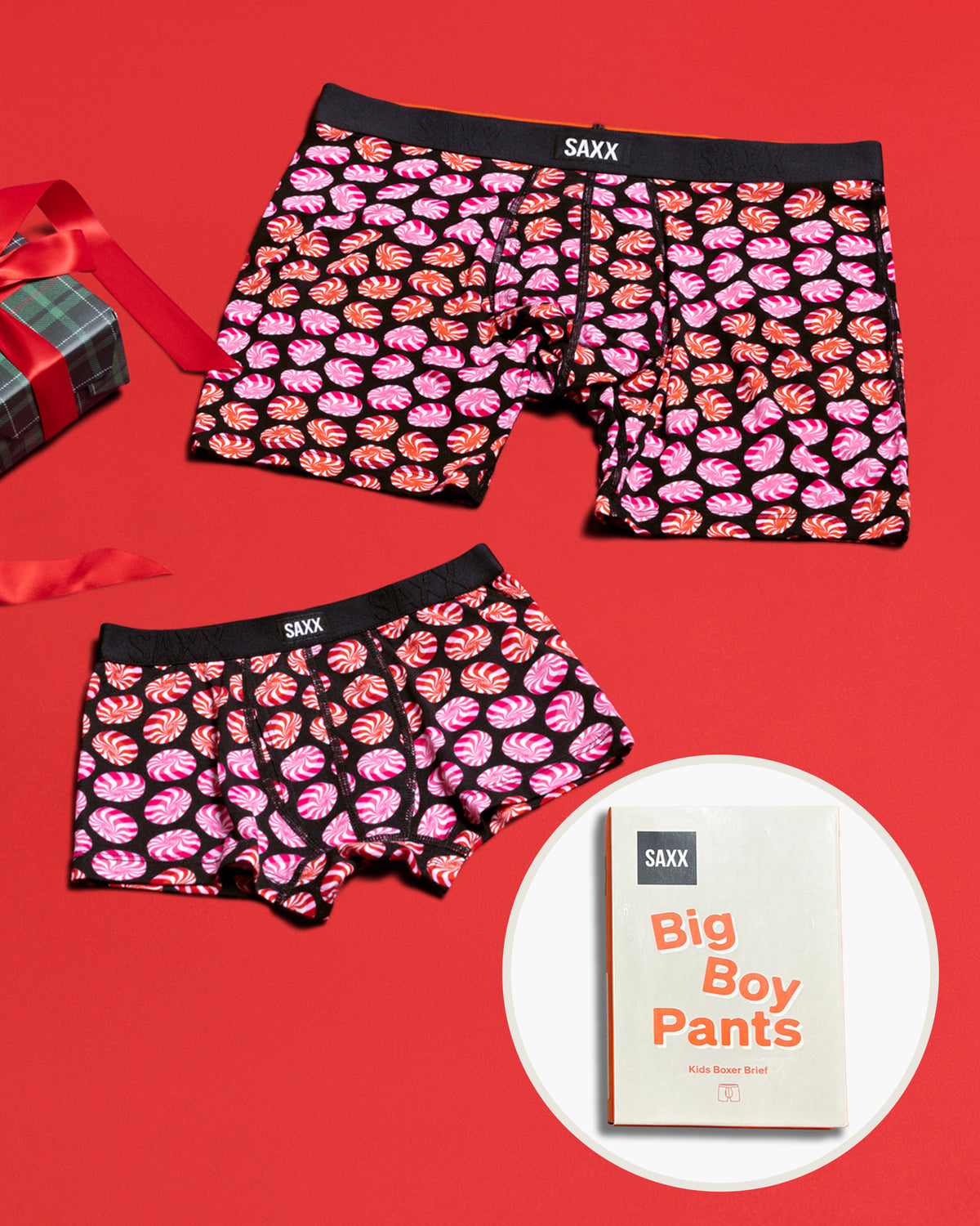 Two pairs of SAXX Father/Son boxer briefs with a candy pattern displayed on a red background with a circular inset showing a box with 'Big Boy Pants'