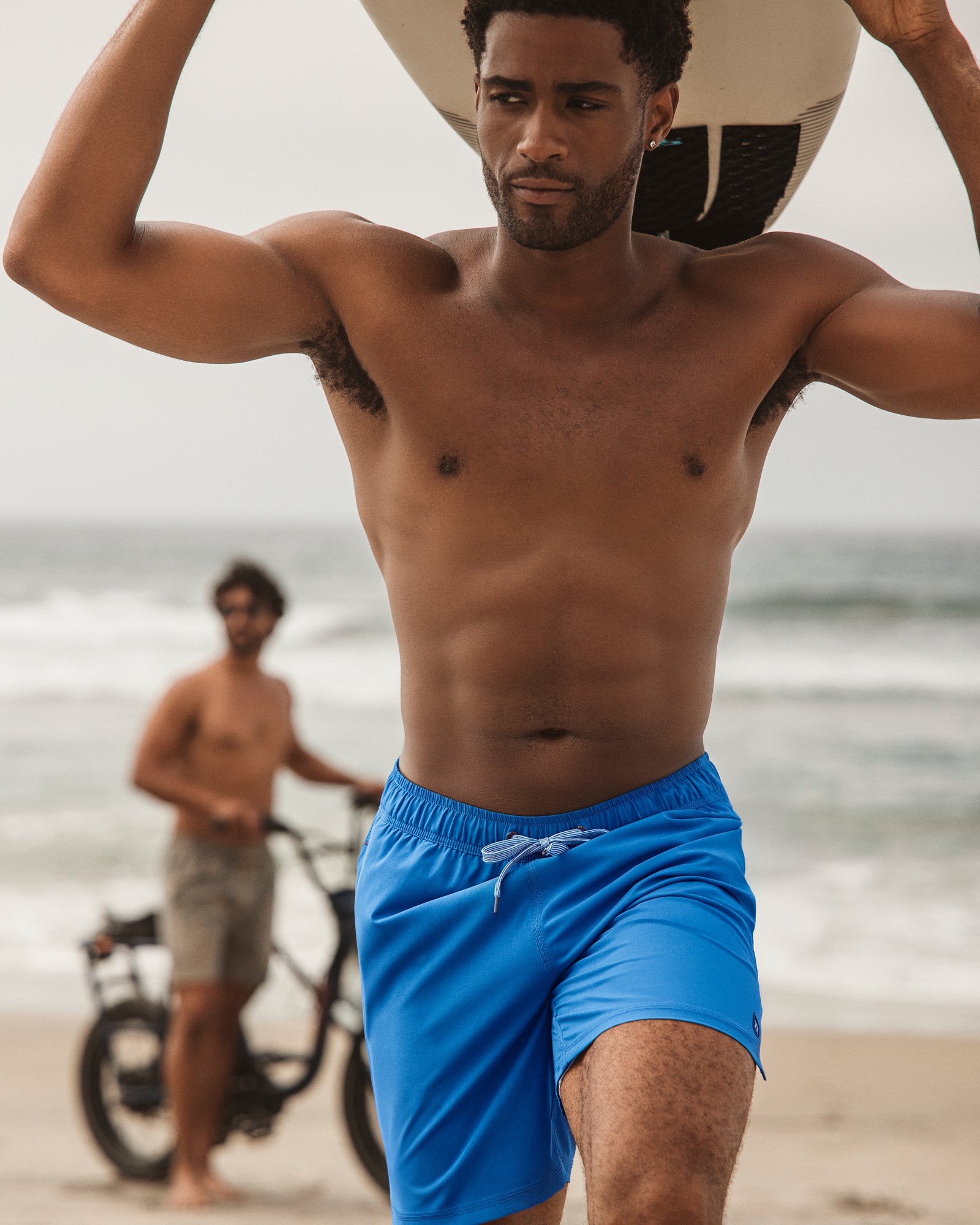 Man in blue swim shorts on a beach with another person and a bicycle in the background.