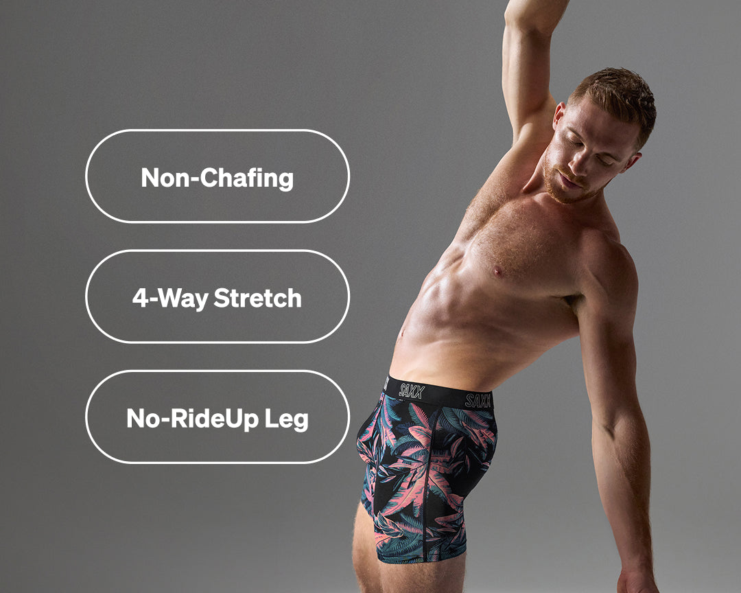 A man wearing patterned boxer briefs poses against a gray background with three white rounded text boxes on the left reading 'Non-Chafing', '4-Way Stretch', and 'No-RideUp Leg'. The underwear has a dark base with pink and teal tropical leaf print and a black waistband with white branding.