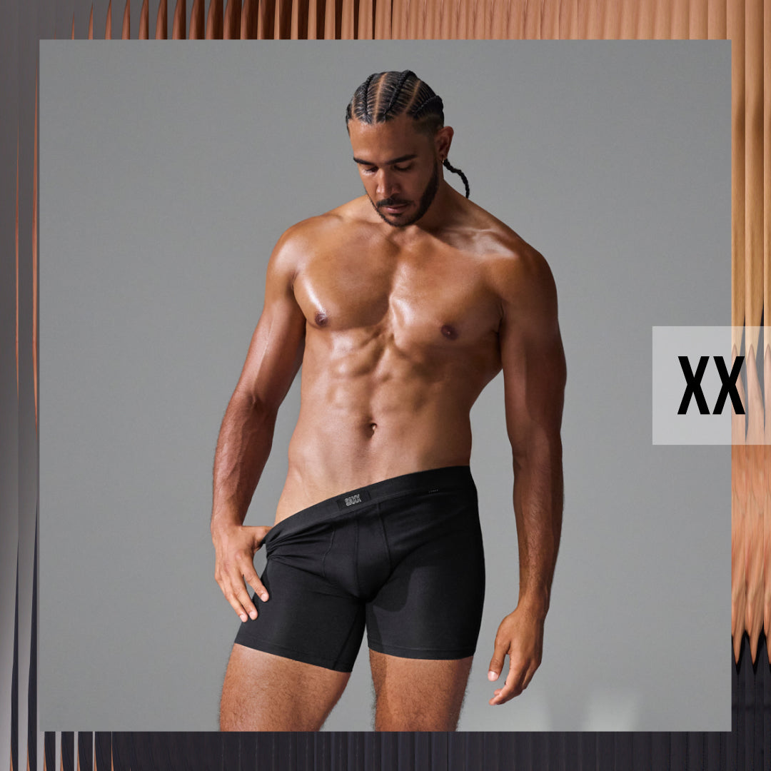 A shirtless male model wearing black boxer briefs, standing against a gray studio background with a wood-textured border and a white 'XX' size label on the right.
