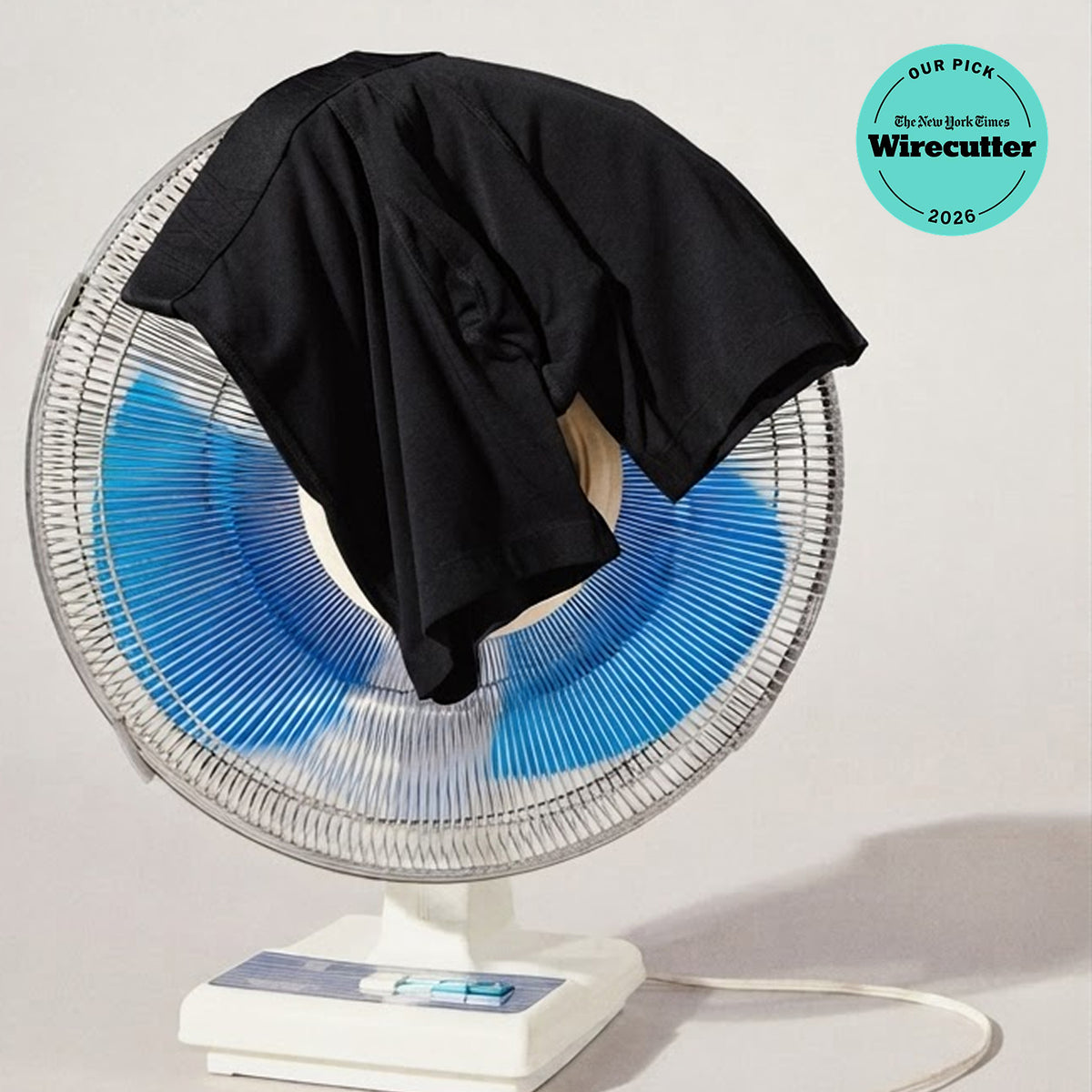 A black short-sleeve T-shirt draped over the front of a white desk fan with blue blades, photographed against a light neutral background. A circular 'Our Pick Wirecutter 2026' badge appears in the top right corner.