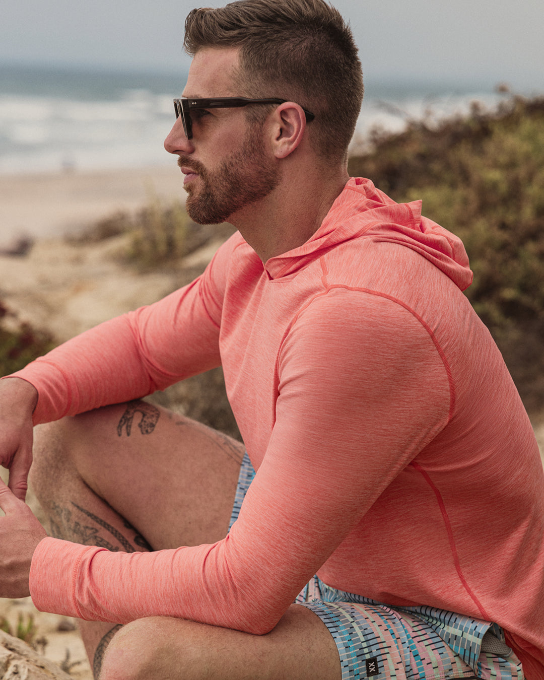 Man wearing a pink hoodie and colorful shorts sitting on a beach.