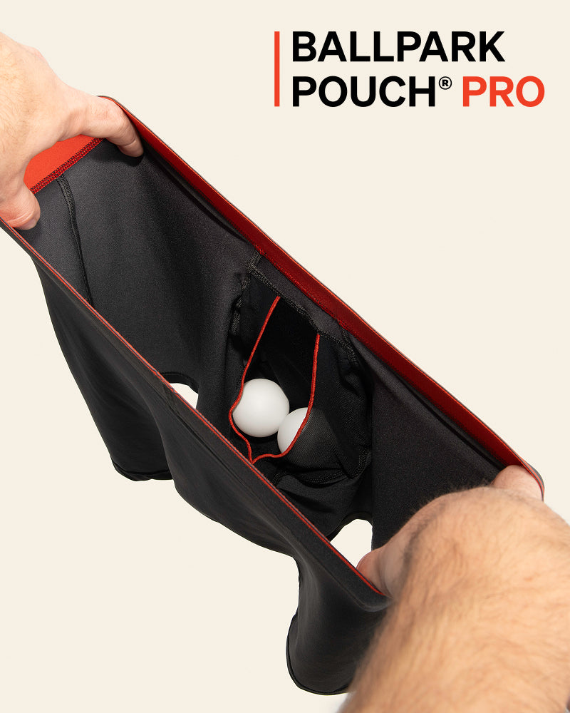 A close-up of a person's hands holding open a black Ballpark Pouch Pro bag with red trim, showing its interior storage compartments and white golf balls inside, against a light beige background with the product logo in the top right.