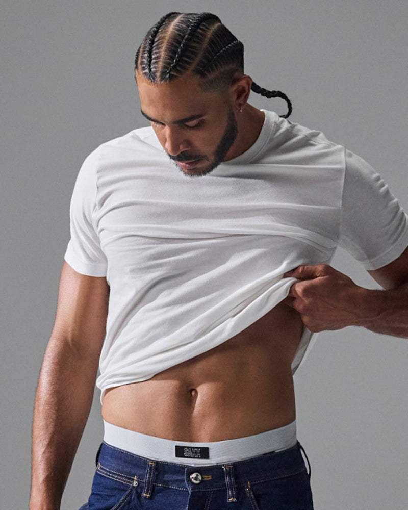 A man wearing a white short-sleeve crewneck t-shirt and blue jeans, lifting the shirt to reveal a white waistband with a black label that reads 'SOME'.