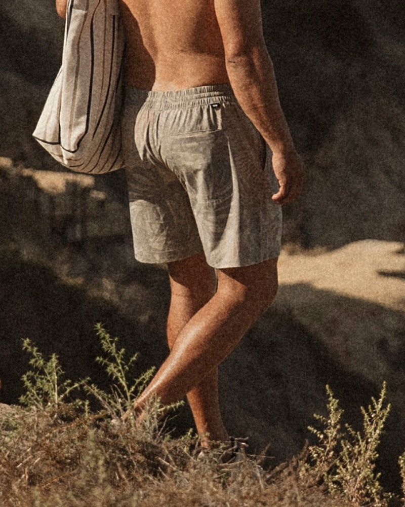 A man from behind wearing light beige linen shorts and carrying a striped canvas tote bag, walking outdoors on dry grass.