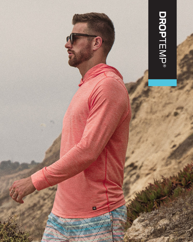 A man wearing a coral pink long-sleeve performance hoodie and patterned shorts walking outdoors on a rocky, sandy hillside. A vertical black Droptemp brand banner with a bright blue accent is on the right side of the image.