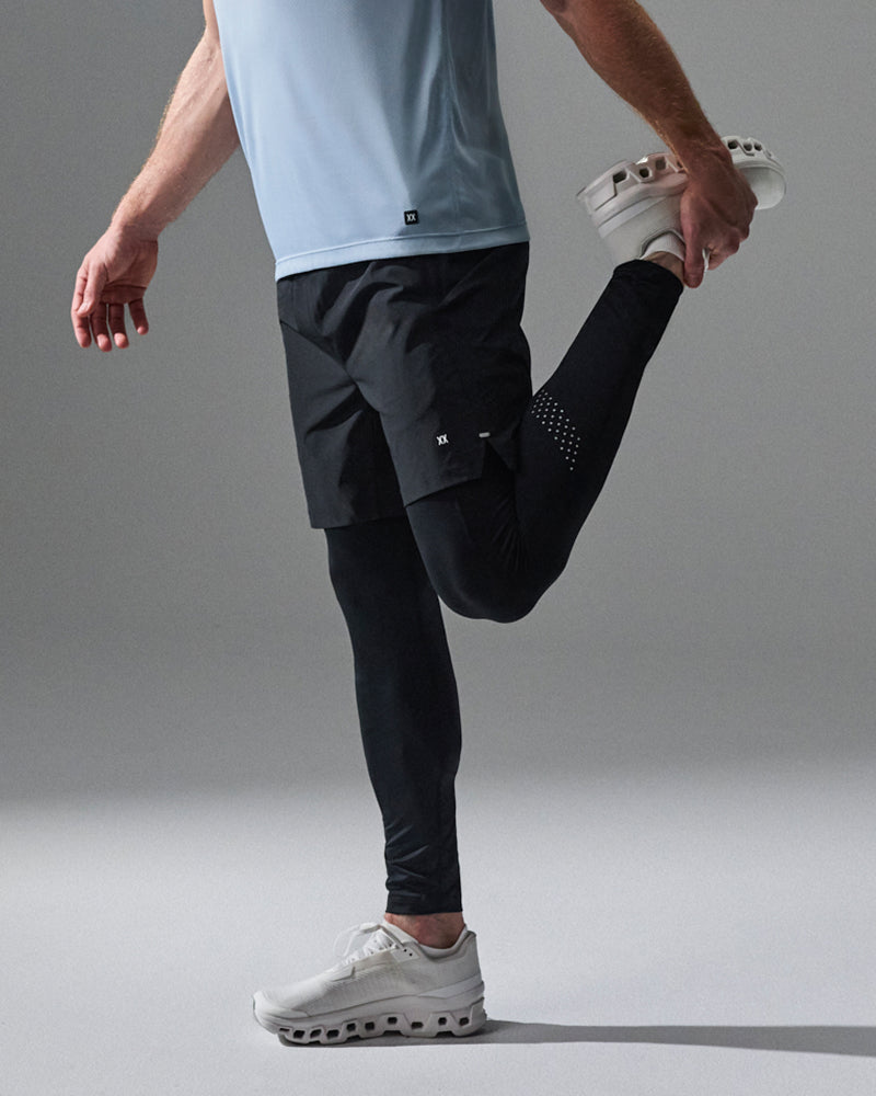 A man in a light blue athletic shirt and black running shorts is performing a leg stretch, holding his foot up. He is wearing black compression leggings and white running shoes against a neutral gray studio background.