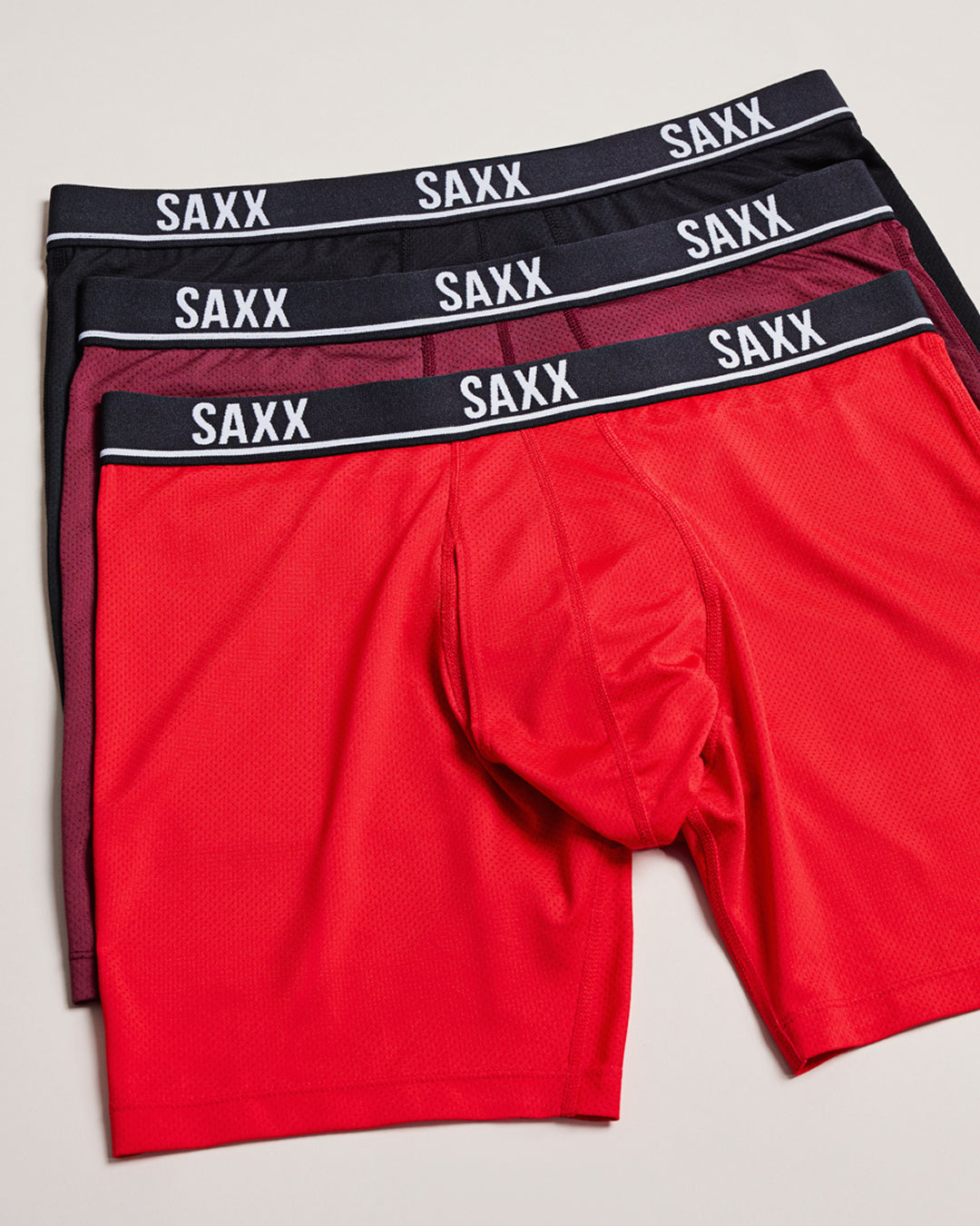 Stacked SAXX boxer briefs in red burgundy and black showing breathable fabric and supportive pouch design
